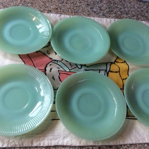 Puede incluir: Seis platillos de vidrio verde con borde acanalado. Los platillos están dispuestos en semicírculo sobre una tela estampada de color blanco y amarillo.