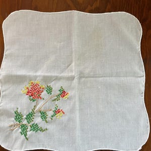 Puede incluir: Pa&ntilde;uelo blanco con borde festoneado y dise&ntilde;o floral de punto de cruz. El bordado presenta flores y hojas rojas, amarillas y verdes sobre un tallo marr&oacute;n. El pa&ntilde;uelo es cuadrado.