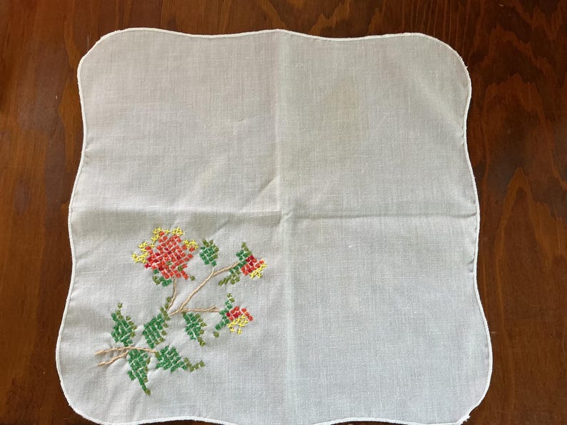 Puede incluir: Pa&ntilde;uelo blanco con borde festoneado y dise&ntilde;o floral de punto de cruz. El bordado presenta flores y hojas rojas, amarillas y verdes sobre un tallo marr&oacute;n. El pa&ntilde;uelo es cuadrado.