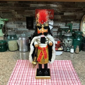 Könnte beinhalten: Ein Nussknacker aus Holz mit schwarzem Bart, roten und goldenen Kleidern und einem weißen, pelzbesetzten Umhang. Der Nussknacker hält ein goldenes Objekt in seiner rechten Hand.