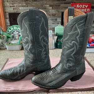 May include: A pair of dark grey cowboy boots featuring a textured snakeskin-patterned leather foot and a smooth leather shaft with decorative white stitching. The boots have a pointed toe, a stacked heel, and pull tabs at the top of the shaft.