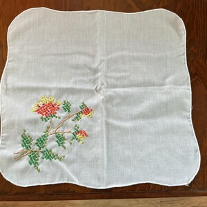 Puede incluir: Pa&ntilde;uelo cuadrado blanco con un dise&ntilde;o floral de punto de cruz. El bordado presenta flores y hojas rojas, amarillas y verdes sobre un tallo marr&oacute;n. Los bordes del pa&ntilde;uelo tienen un dise&ntilde;o festoneado.