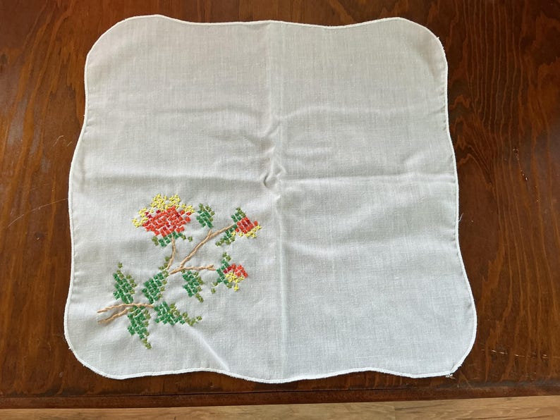 Puede incluir: Pa&ntilde;uelo cuadrado blanco con un dise&ntilde;o floral de punto de cruz. El bordado presenta flores y hojas rojas, amarillas y verdes sobre un tallo marr&oacute;n. Los bordes del pa&ntilde;uelo tienen un dise&ntilde;o festoneado.