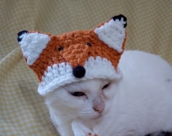 Könnte beinhalten: Eine weiße Katze trägt eine gehäkelte orangefarbene und weiße Fuchs-Mütze. Die Katze liegt auf einer gelben und weißen karierten Oberfläche mit braunen Papier-Serpentinen.