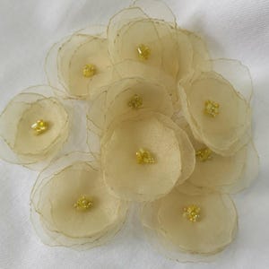 Peut inclure: Un ensemble de fleurs en tissu jaune clair, composées de plusieurs couches et d'un centre perlé. Les pétales sont translucides. Les fleurs sont disposées en groupe. Idéales pour l'artisanat ou les embellissements, ajoutant une touche d'élégance.