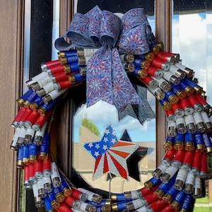 May include: A patriotic wreath made from red, white, and blue shotgun shells. The wreath is adorned with a large bow and a star-shaped decoration, creating a festive and decorative piece for the front door or wall.