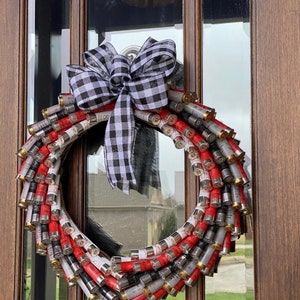 May include: A decorative wreath made from red, white, and gray shotgun shells, accented with a black and white checkered bow. The wreath is displayed on a wooden door with glass panels, creating a rustic and unique home decor item.