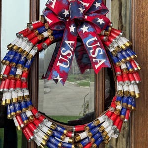 May include: A patriotic wreath made from red, white, and blue shotgun shells. The wreath is adorned with a large bow featuring the text "USA" and white stars on a red and blue background. This decorative wreath is perfect for celebrating Independence Day.
