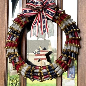 May include: A patriotic wreath made from red, white, and blue shotgun shells. The wreath is adorned with a large bow featuring stars and stripes. A wooden star with an American flag design is placed in the center.