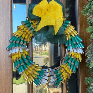 May include: A green and yellow wreath made of shotgun shells with a yellow and green burlap bow.