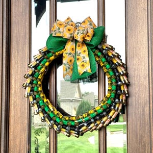 May include: A handmade wreath made of shotgun shell casings in yellow, green, and black, with a sunflower-patterned bow and green burlap ribbon. The wreath is hanging on a wooden door.