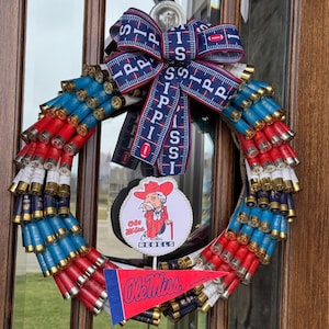 May include: A red, white, and blue wreath made of shotgun shells with a blue and white ribbon bow and a Ole Miss Rebels sign in the center.