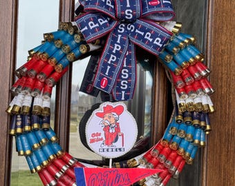 May include: A red, white, and blue wreath made of shotgun shells with a blue and white ribbon bow and a Ole Miss Rebels sign in the center.