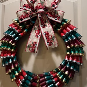May include: A festive Christmas wreath crafted from red, green, and gold shotgun shells. A large bow with a beige background and red trucks carrying Christmas trees adorns the top. This unique wreath is a decorative holiday accent.