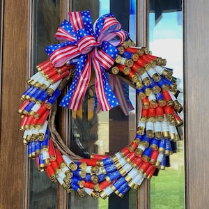 May include: A patriotic wreath made from red, white, and blue shotgun shells. The shells are arranged in a circular pattern around a natural twig base. A large bow with red, white, and blue striped and star-spangled ribbons adorns the top of the wreath.