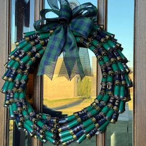 May include: A decorative wreath made from green and navy blue shotgun shells. The wreath is adorned with a large plaid bow in shades of green and navy blue. The wreath is hanging on a glass door.