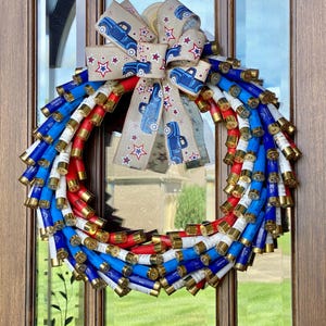 May include: A red, white, and blue patriotic wreath made of shotgun shells with a burlap bow featuring a blue truck and stars.