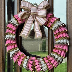 May include: A pink and white wreath made of shotgun shells with a pink and white striped bow.