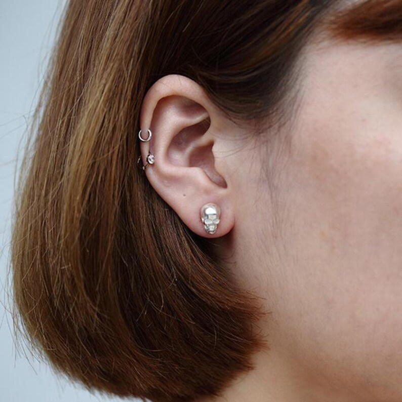 Pu&ograve; includere: Primo piano di un orecchio con un orecchino a forma di teschio in argento. L'orecchino &egrave; piccolo e dettagliato, con una superficie testurizzata.