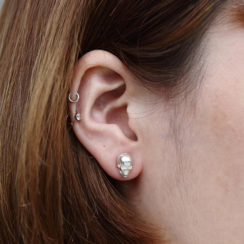 Pu&ograve; includere: Primo piano di un orecchio con un orecchino a forma di teschio in argento. L'orecchino &egrave; piccolo e dettagliato, con una superficie testurizzata.