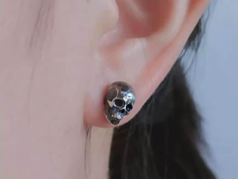 Pu&ograve; includere: Un orecchino a forma di teschio in argento con un design dettagliato. L'orecchino &egrave; indossato sul lobo dell'orecchio sinistro.