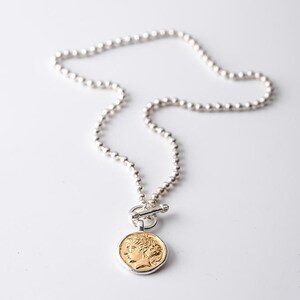 May include: A silver chain necklace with a gold coin pendant featuring a profile of a person. The chain is made of small, round beads.