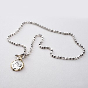 May include: A silver chain necklace with a silver and gold coin pendant featuring a profile of a woman. The chain is made of small, round beads.