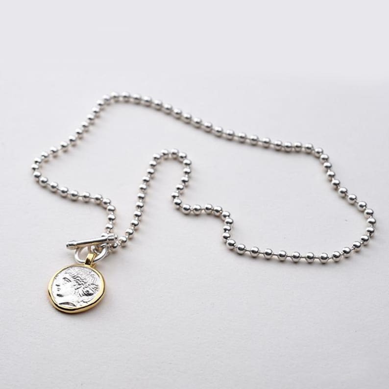 May include: A silver chain necklace with a silver and gold coin pendant featuring a profile of a woman. The chain is made of small, round beads.