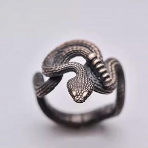 Könnte beinhalten: Ein silberner Ring mit einem detaillierten Schlangendesign. Die Schlange ist zusammengerollt und hat eine strukturierte Oberfläche. Der Ring ist ein Statement-Piece und wäre ein einzigartiges Geschenk.