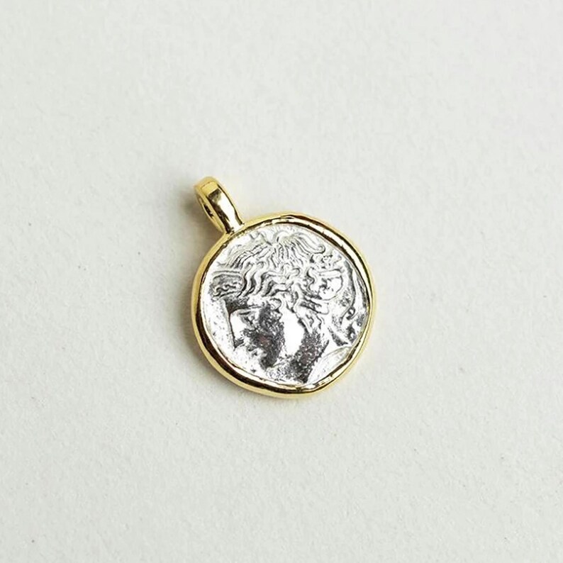 May include: A gold-toned pendant with a silver-toned coin featuring a profile of a person with a crown. The coin is surrounded by a gold-toned rim.