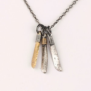 Puede incluir: Un collar de cadena de plata con tres colgantes de cuchillo de plata y oro. Los cuchillos son de diferentes tamaños y tienen un acabado texturizado.