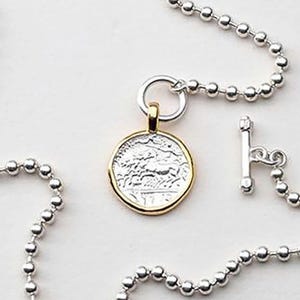 May include: A silver chain necklace with a gold-rimmed silver coin pendant featuring an engraved design. The necklace has a toggle clasp.