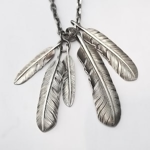 May include: A silver necklace with a pendant featuring five detailed feather charms. The feathers are intricately designed with a textured surface and appear to be made of metal.