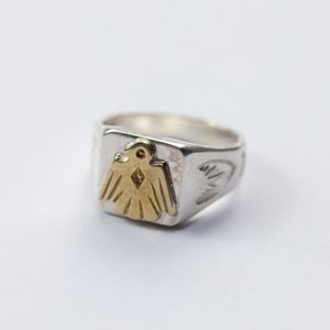 Op de afbeelding: Een zilveren ring met een gouden adelaarontwerp op de bovenkant. De ring heeft een vierkante vorm en is aan de zijkanten versierd met een patroon.