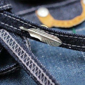 May include: A silver feather charm on a dark blue denim background. The feather charm is detailed and has a textured surface.