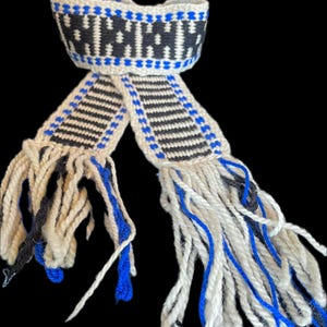 May include: A handwoven scarf with a geometric pattern in black, white, and blue. The scarf features a band of black and white design at the top, with stripes and fringe at the ends. The fringe is a mix of white, black, and blue yarn.