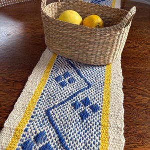 May include: A woven basket with two lemons sits on a blue and white patterned table runner with yellow borders. The basket is made of natural fibers and has two handles. The runner is on a wooden table.