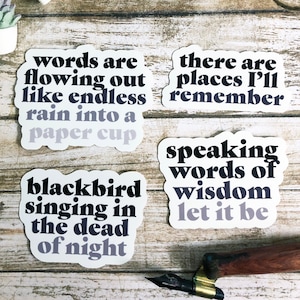 Könnte beinhalten: Vier Aufkleber mit schwarzem Text auf weißem Hintergrund mit einem violetten Farbverlauf. Die Aufkleber zeigen folgenden Text: "words are flowing out like endless rain into a paper cup", "there are places I'll remember", "blackbird singing in the dead of night", und "speaking words of wisdom let it be".