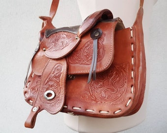 May include: A brown leather saddle-shaped crossbody bag with intricate floral tooling and white stitching. The bag has a flap closure and a long strap.