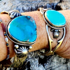 May include: Two silver-toned bracelets with large, oval turquoise stones. The bracelets have intricate metalwork and are worn on a wrist. A beaded bracelet is also visible. The background is a blurred, natural setting.