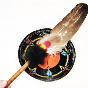 May include: A Native American style feather fan with a black, circular base. The fan features a large brown and white feather, a black fluffy section, and a wooden handle. The base has colorful geometric designs.