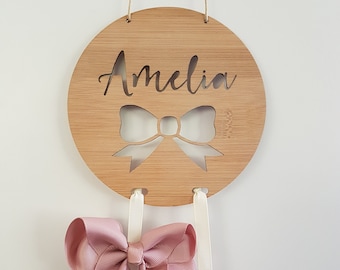 May include: A round wooden sign with the name "Amelia" cut out and a bow shape in the center. Two pink bows are hanging from the sign on white ribbons.