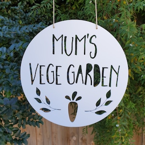 May include: White round wooden sign with the text "Mum's Vege Garden" in black lettering. The sign has a carrot and two leafy branches cut out of the wood.