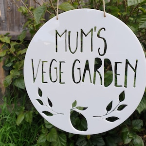 May include: White circular sign with the text "Mum's Vege Garden" in black lettering. The sign has a cut-out design of leaves and a lemon.