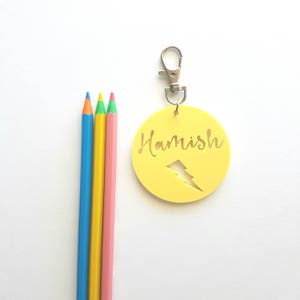 May include: Yellow round keychain with the name "Hamish" and a lightning bolt cutout. The keychain has a silver clip. Three colored pencils are in the background.