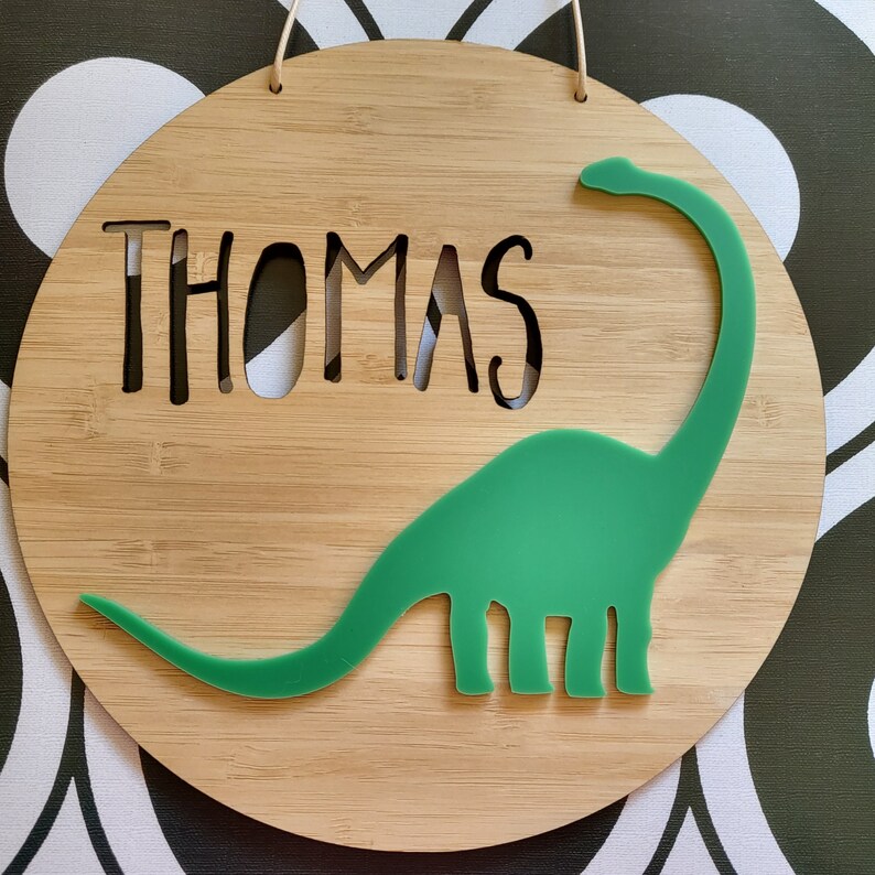 May include: A wooden sign with the name "Thomas" cut out in the center. A green dinosaur silhouette is attached to the sign.