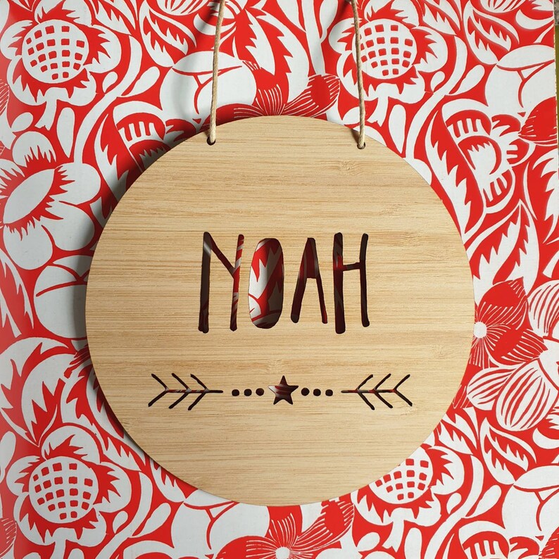 May include: A round wooden sign with the name "Noah" cut out in the center. The sign has a decorative arrow design and a star at the bottom.
