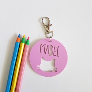 May include: A pink round keychain with a cat silhouette cut out and the name "Mabel" written in white. The keychain has a silver metal clasp.