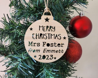 May include: A wooden Christmas ornament with the text "Merry Christmas* Mrs Foster from Emmett *2023*" in a white font. The ornament is hanging on a green Christmas tree.