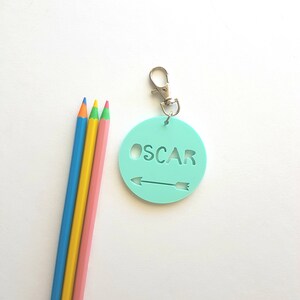 May include: A light blue round keychain with the name "OSCAR" cut out and an arrow pointing to the right. The keychain is attached to a silver split ring.